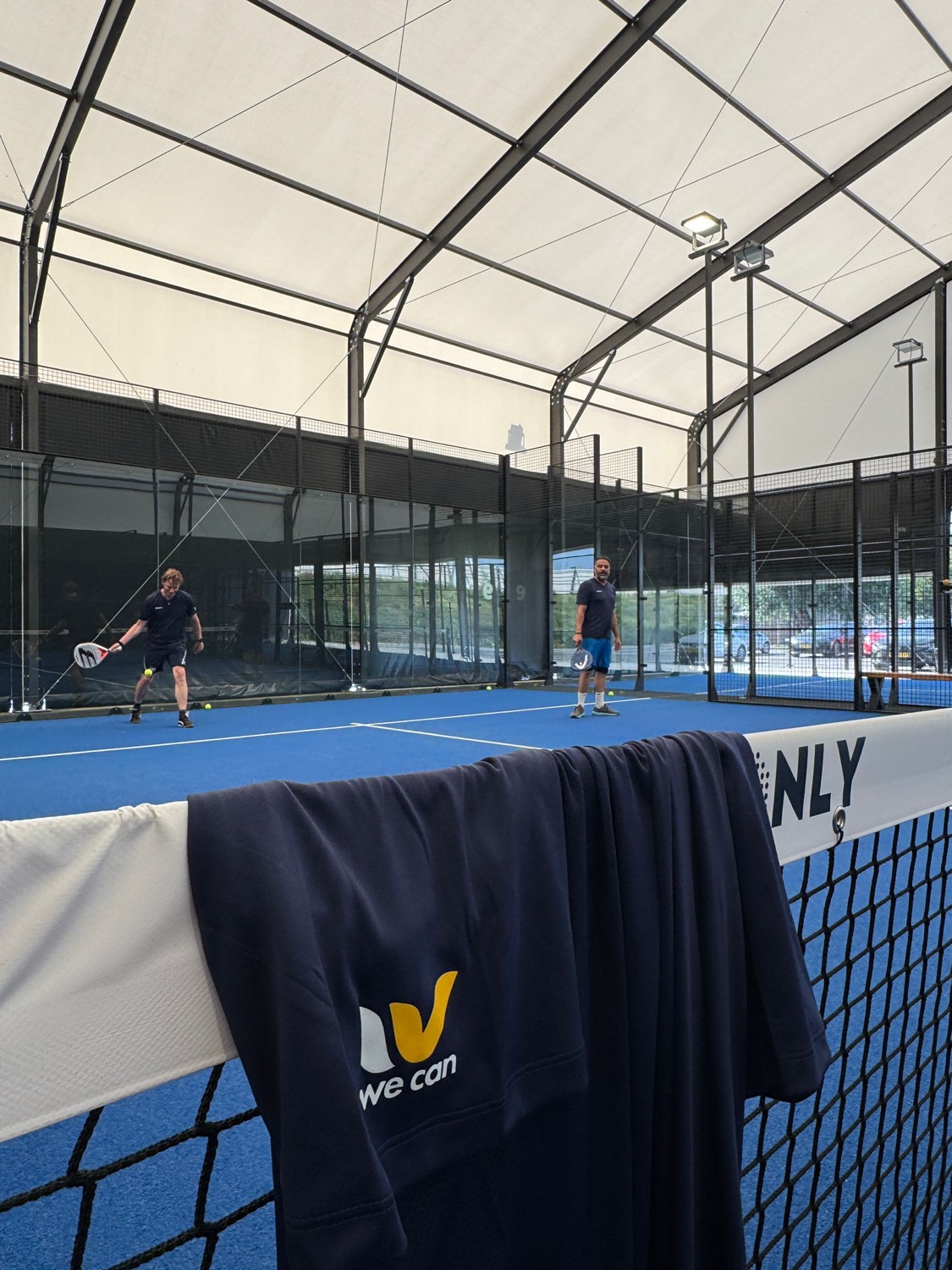 Players on the padel court at We Can's first Monthly Padel, with a We Can towel over the net